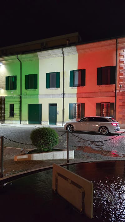 The image depicts a street scene at night in Peschiera del Garda, Italy. A white Audi station wagon is parked on a cobblestone street in front of a building whose facade is painted with the colors of the Italian flag: green on the left, yellow in the middle, and red on the right. The building has several windows with green shutters, and some of the windows are illuminated from within. A neatly trimmed green bush is situated in the foreground, between the street and a shallow water feature or fountain. The ground is wet, suggesting it has recently rained, and reflections of the red light from the building can be seen on the pavement and in the water. The overall atmosphere is dark, with artificial lighting highlighting the building and the car.