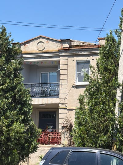 A two-story building with a beige facade is visible, partially obscured by green trees. The building features a central balcony on the upper floor with a black metal railing, and a smaller red metal balcony on the lower floor. There is a dark blue door on the ground floor. A circular decorative element is present above the upper balcony, and colorful triangular flags are strung across it. A window on the upper right side of the building displays a sticker of a fish with the text "SMILE FISH". The roofline is uneven and appears to be in a state of disrepair. A dark colored car is parked in front of the building. The sky above is clear and blue, with power lines crisscrossing the scene. This image was captured on Gorky Street in Taganrog, Russia.