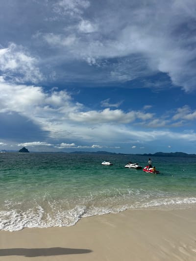 A bright, sunny day with scattered clouds over a turquoise ocean. The foreground shows a sandy beach with gentle waves lapping at the shore, casting a long shadow on the sand. In the middle ground, several jet skis are visible on the water. Two jet skis are white and black, one with a person standing on it. A red and white jet ski is to the right, with two individuals on it. In the distance, lush green islands form a mountainous backdrop against the horizon. The overall scene depicts a tropical, recreational setting in Phru Nai, Thailand.