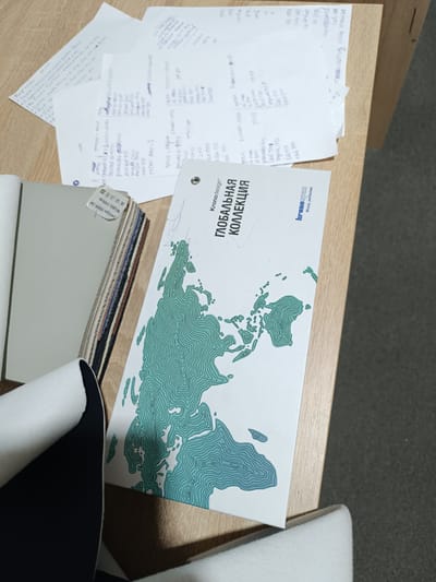 The image shows a close-up view of a desk with several items related to interior design or material samples. The primary subjects are samples of materials, likely for furniture or flooring, stacked in a binder. There is also a brochure with a stylized map of the world on it, featuring a green contour-line design. Scattered papers with handwritten notes and lists of items, possibly prices or codes, are also visible. The setting appears to be an office or workspace.
The activity taking place seems to be related to design selection or project planning, with the materials and brochure laid out for review. There are no people visible in the image. The lighting suggests it is daytime.
Notable details include text on the material samples and the brochure. One tag reads "Design:0009-3# Width: 140cm". The brochure has text in Russian, including "Kronodesign", "ГЛОБАЛЬНАЯ КОЛЛЕКЦИЯ" (Global Collection), and a logo with the text "kronospam Wood, perfected". The handwritten notes on the papers contain a mix of Cyrillic and Latin characters, with what appear to be names and numerical values. One section of notes is labeled "Кофейни" (Coffee shops) and lists various coffee drinks with associated numbers. Another list contains items like "Лисиков", "KALIA", and "дресата" with prices. The overall impression is one of organization and selection within a professional context.