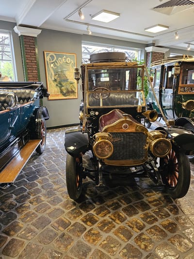 The image shows an interior view of a museum or exhibition hall, likely featuring vintage automobiles. The location is possibly Архангельское, Russia, inferred from the presence of vintage cars, which is consistent with museum or historical displays.
The foreground is dominated by an antique car with a black chassis, brass or bronze-colored accents, and large, classic headlights. The car has a radiator with a black mesh grille. Its tires are thin and likely made of solid rubber.
In the background, other vintage cars are partially visible. There's a large poster of a Peugeot car on a wall, framed and hung above a faux brick wall.
The floor is paved with roughly textured stones, creating a vintage ambiance. The lighting is provided by modern fixtures on the ceiling, creating an artificial illumination environment. The setting suggests a controlled, museum-like environment for displaying and preserving these historical vehicles. There are no people visible in the frame.