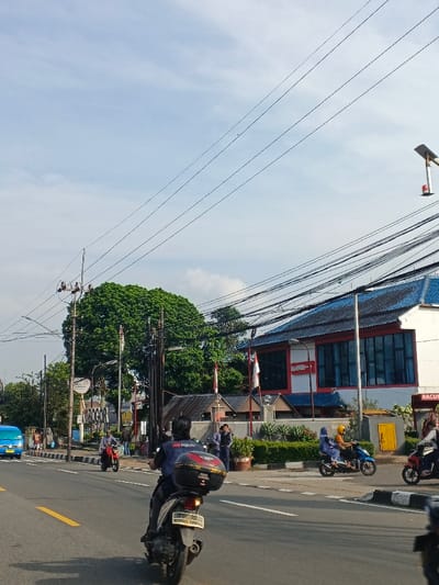 A street scene in Bogor, Indonesia. Several people on motorbikes drive along a road marked with yellow lines. A sign reading "Bogor" is visible beside a large tree. Buildings line the street, one with a blue roof and red trim, and another with "RACUN" written on the sign. Power lines crisscross overhead against a backdrop of a blue sky with scattered clouds.