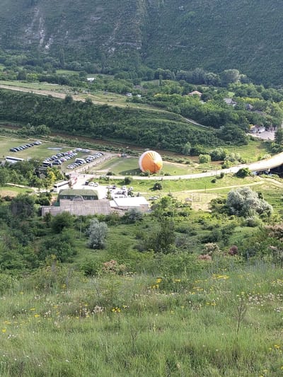 The image captures a rural landscape near Brănești, Moldova, characterized by lush green vegetation, rolling hills, and a winding road. In the foreground, a grassy field dotted with wildflowers, including yellow dandelions and white seed heads, stretches towards the middle ground.

Dominating the middle ground is a large, partially inflated hot air balloon, predominantly orange and white, situated on a paved area near a road. Adjacent to the balloon is a complex of buildings with green roofs, possibly a recreational facility or a local establishment. Several vehicles, including cars and what appears to be a bus, are parked in a lot behind these buildings.

Further into the scene, a river meanders through the landscape, bordered by dense trees. A yellow bridge arches over the river, connecting two sides of the area. The background is filled with a densely forested hillside, displaying various shades of green and textured foliage, with a rocky cliff face visible on the far left. The sky is not clearly visible due to the angle of the shot and the overhead canopy of trees and hills. There are no people clearly depicted in the scene, though the presence of vehicles suggests potential human activity. The overall impression is of a serene, natural environment with elements of human infrastructure.