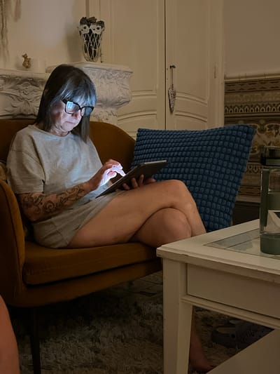 A seated woman with short, gray hair and glasses is looking at a digital tablet, which is being held in her hands and the bright light from the screen illuminates her face. The woman has a tattoo on her left arm and she is wearing a gray t-shirt. She is sitting in a mustard-colored chair with a blue pillow behind her, situated at a table with a white surface. The floor appears to be carpeted with a neutral gray carpet. In the background, there is a wall with decorative trim, including a floral arrangement and a decorative fixture. There is no specific evidence to indicate the location of Montélimar, France.