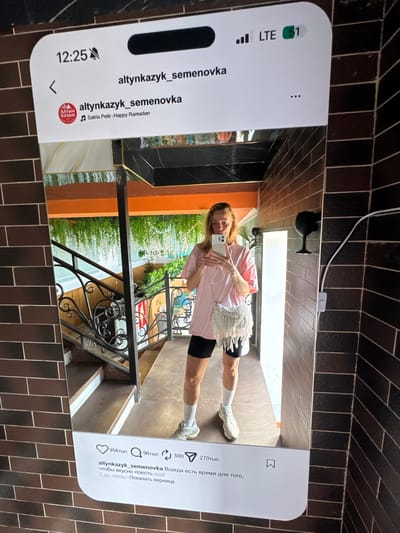 A medium shot depicts a person holding an iPhone, which displays an Instagram page with the username "altynkazyk_semenovka". The person is positioned in a building with a staircase, a metallic railing, and an orange wall visible in the background. There is a glimpse of greenery through the windows. The background, including the wall and the surface where the phone is placed, is tiled with dark brown rectangular tiles. A white wire runs along the upper part of the image.