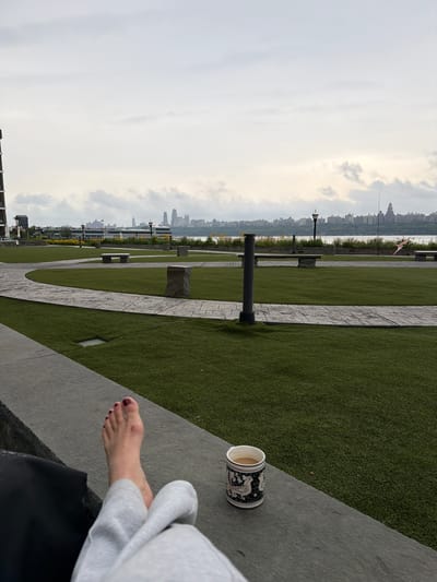 The image depicts a peaceful outdoor scene in Edgewater, United States of America. In the foreground, a person's feet, adorned with dark red nail polish, rest on a dark stone ledge. They are wearing light gray sweatpants. To the right of their feet, a white ceramic mug with a dark blue decorative pattern sits on the ledge, filled with a light brown beverage.

The background reveals a meticulously maintained grassy area with curved stone pathways. Several stone benches and decorative stone pillars are strategically placed within the green space. Beyond the park, a body of water is visible, with a city skyline stretching across the horizon under a cloudy sky. A few buildings with distinct architectural features, including taller structures, are discernible within the distant cityscape. A kite, shaped like a diamond and colored pink and white, flies on a thin string in the middle distance. The overall atmosphere is serene and relaxing.