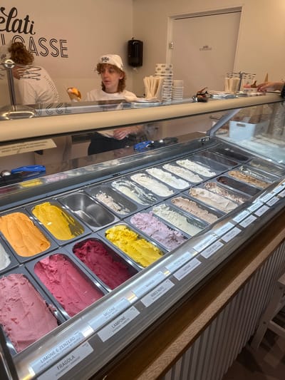 The image captures the interior of a brightly lit gelato shop, "Gelati Schulgasse," located in Winterthur, Switzerland. The primary focus is a long, glass-fronted display counter filled with numerous metal trays of colorful gelato and sorbet.
Above each flavor, labels provide names in Italian, German, and often English. Visible flavors include "LIMONE E ZENZERO (LEMON WITH GINGER)," "FRAGOLA (STRAWBERRY)," "MELONE (MELON)," "LAMPONE (RASPBERRY)," "MANGO," "RIBES NERO (BLACK CURRANT)," "PISTACCHIO (PISTACHIO)," "SAFRAN," "BIRCHERMÜESLI," "VANIGLIA (VANILLA)," "YOGURT MORA (BLACKBERRY YOGURT)," "HONIG," "YOGURT NATURA," "FIOR DI LATTE," "NOCCIOLA," "BANOFFEE PIE," "CHAI LATTE," and "MOHNGCHEN." Several fruit flavors are explicitly marked as "SORBET."
A young staff member with curly hair, wearing a white "Gelati" cap and shirt, stands behind the counter, holding an ice cream cone and looking towards the viewer. Their reflection and the shop's wall text, "Gelati Schulgasse," are visible on the counter's glass. Another person's hand is partially visible at the top edge of the frame, possibly reaching for cones or serving.
The counter features stacked white paper cups with the "Gelati" logo and wrapped cutlery. White walls in the background display large black text spelling out "Gelati SCHULGASSE" and a partially obscured "Gelati GASSE." A sign on the counter warns, "ALLERGIEN UND HERKUNFTS INFORMATIONEN WENDET EUCH BITTE AN DAS PERSONAL" (Allergy and origin information, please contact staff). The scene depicts a typical, well-lit operation within a gelato shop, suggesting a regular business day.