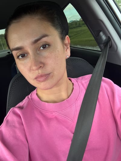 A young woman with dark hair pulled back and wearing a pink sweatshirt is seated in a car, wearing a seatbelt. She has freckles and is looking towards the camera. The background outside the car window shows a rural landscape with green fields and a cloudy sky. The location is identified as Verkhneyarkeyevo, Russia.