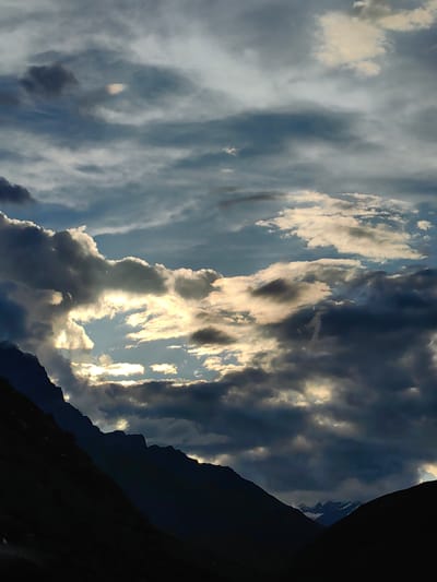 The image depicts a dramatic sky filled with heavy, dark clouds interspersed with patches of bright sunlight breaking through. The foreground is dominated by the dark silhouettes of steep mountain slopes, likely part of the Himalayan range. In the distance, faint outlines of snow-capped peaks are visible through a gap in the clouds. The overall impression is one of rugged, natural beauty, with the interplay of light and shadow creating a powerful atmosphere. The location is identified as Damphug, India. There are no people, animals, or man-made structures clearly visible in the image.