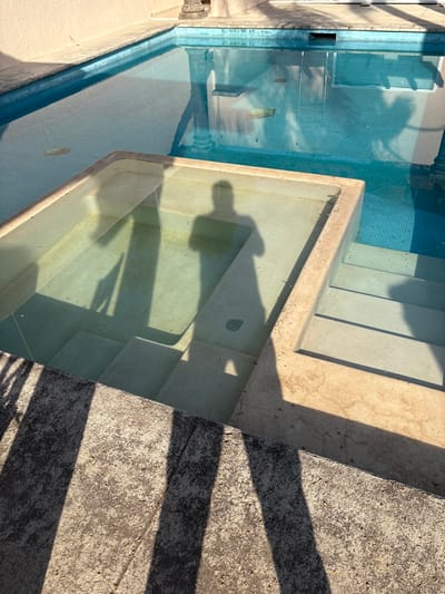 This image depicts an outdoor swimming pool area located in Chamilpa, Mexico. In the foreground, a textured, light-colored concrete surface is visible, marked by several dark, elongated shadows. The mid-ground features a rectangular, stepped pool structure, likely a spa or shallow entry, filled with clear water, allowing its internal concrete steps to be seen. A distinct human-shaped shadow, consistent with a person holding a camera or phone, is cast into this stepped pool and onto its internal surfaces. To the right of this, a larger swimming pool with blue mosaic tiling extends into the background. The main pool also contains clear water, reflecting portions of surrounding architecture, including what appears to be a stone column. A light-colored, irregularly shaped object floats on the surface or rests near the bottom of the main pool. The background consists of a light-toned wall, behind which part of a decorative stone pillar is visible. The scene is illuminated by direct sunlight, producing sharp, contrasting shadows.