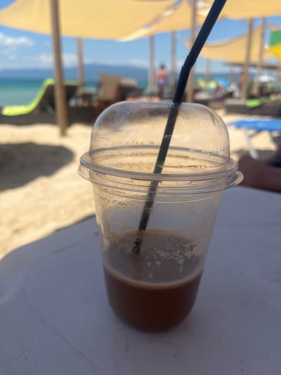 A close-up shot focuses on a nearly empty plastic cup with a domed lid and a black straw, resting on a white surface. The cup contains a small amount of brown liquid with foam residue on the sides. The background is a blurred beach scene, likely in Ofrynio Beach, Greece, under a bright, sunny sky. Beach umbrellas, lounge chairs, and the sea are visible. The image captures a moment of leisure on a warm day.