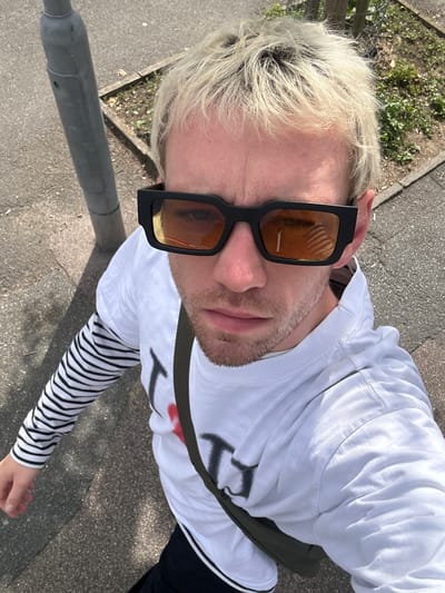 In a high-angle, self-taken photograph, a person with bleached blonde hair, wearing black-rimmed sunglasses with amber lenses, stands on a paved surface in Folkestone and Hythe District, United Kingdom. The individual is dressed in a white long-sleeved t-shirt layered over a striped long-sleeved shirt with black and white horizontal stripes. A dark green strap, possibly from a bag, is visible across the chest. The background consists of a grey concrete sidewalk, a patch of soil with sparse vegetation, and a grey metal pole, likely a lamppost. The lighting suggests it is daytime, with the sun casting shadows. The image captures a close-up view of the person's face and upper body, with the environment occupying the periphery.