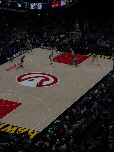 The image shows a basketball game in progress at the State Farm Arena in Atlanta, United States. The teams are wearing red and green uniforms. The center of the court features the Atlanta Hawks logo. Spectators are in the surrounding seats. The game clock shows 5:34 in the 1st quarter, with a score of 12 to 6. Signage for Ticketmaster, ks.com, Kia, and Tissot can be seen around the court.