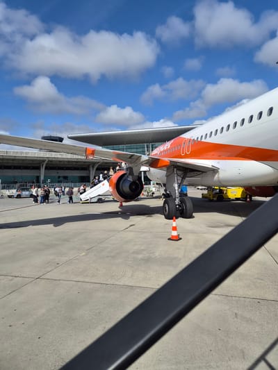 The image shows the side of an EasyJet Airbus A320 parked at a gate, likely at Francisco Sá Carneiro Airport (OPO) near Moreira, Portugal, given the context. Passengers are disembarking using an air stair, and the jet engine is visible. A yellow ground support vehicle is near the aircraft's landing gear, which has a safety cone placed near it. The sky is visible, with scattered clouds. Part of a fence or railing is in the foreground.