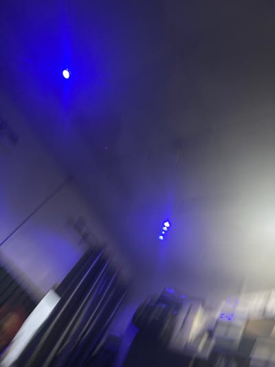 The image depicts an interior scene with motion blur, primarily featuring a predominantly dark ceiling illuminated by several blue lights. One prominent blue LED light is visible in the upper left portion of the frame, appearing as a bright, circular point source with a halo. Further down and to the right, a cluster of four smaller blue LED lights is arranged in a diagonal line. The overall ambiance is dark, with a gradual transition to lighter tones, possibly indicating a wall or a different area of the room. The blur suggests movement during the capture of the photograph, making specific details of objects or people indistinct. There is no visible indication of the location being in Naraguta Hausawa, Nigeria.