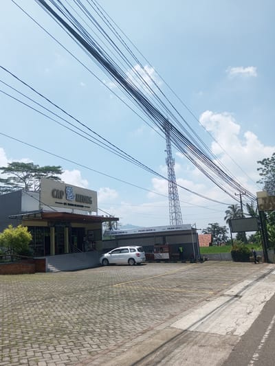 The image shows a retail building with "CAP MEONG" signage under a bright sky with numerous overhead power lines. The building features a storefront entrance with steps leading up, and a parking lot paved with grey interlocking bricks. A white car is parked in one of the spaces. Adjacent to the building is an open-air structure, seemingly a "Pojok UMKM" space. A tall telecommunications tower rises in the background. A figure is visible walking along the road in the lower right. No specific indications point definitively to Nawng Hkar, Myanmar.