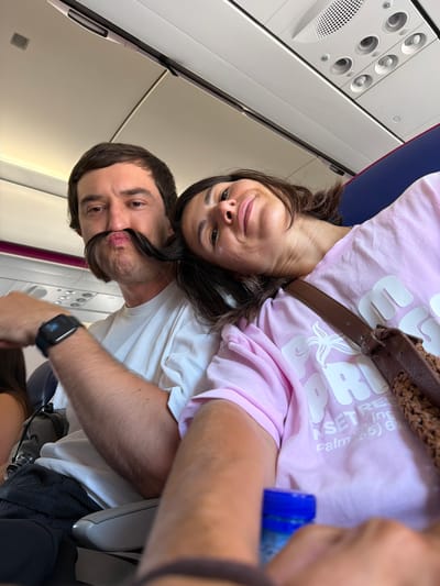 A couple is seated on an airplane, with the man in the foreground and the woman leaning her head on his shoulder. The man has a large, dark mustache made from his own hair. He is wearing a white t-shirt and a smartwatch on his left wrist. The woman is wearing a pink t-shirt and has a brown bag slung over her shoulder. The interior of the airplane cabin is visible, including overhead compartments and seats. The image appears to be a selfie taken by the couple. The location is not identifiable as Donja Cijevna, Montenegro.