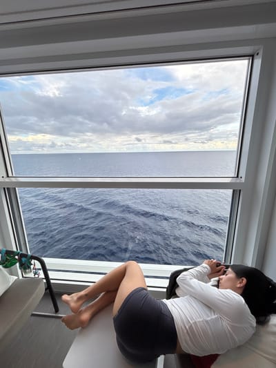 A female individual is observed resting inside a modern cabin or room, likely on a vessel, overlooking the open ocean near the Berry Islands, Bahamas. She is lying on her right side with her back partially turned towards a large, rectangular window framed in white, which dominates the scene. She wears a white long-sleeved top with textured detailing and dark gray shorts. Her dark hair is spread behind her head, and her hands are clasped near her face, which is turned towards a light-colored surface, possibly a pillow. Her bare feet and legs, bent at the knees, are visible. To the left, a portion of a light gray sofa or bench is seen, alongside a black metal frame from which green and red fabric straps are suspended. Through the window, the expanse of the dark blue sea with visible waves extends to the horizon, meeting an overcast sky with patches of lighter clouds.