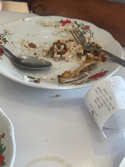 The image depicts a close-up, overhead view of a white, floral-patterned plate on a white table, with remnants of a meal. A fork and spoon are positioned on the plate, amidst scattered white rice, pieces of fried chicken bone, and some green vegetables. A crumpled receipt lies on the table to the right of the plate. The receipt displays text in Indonesian, including the name "Nekan Lodeh", a phone number "082312318809", an address "Ruko Boulevard no.23, Jl Gg Luh Timur, Karawang", and a date and time "Selasa, 26 Jun 2024 13:38". The presence of a receipt from Karawang suggests the location is likely within that city or its vicinity, which is in West Java, Indonesia. A portion of a person's arm is visible in the upper right background, suggesting someone was sitting at the table. The overall scene captures the aftermath of a meal at a restaurant.