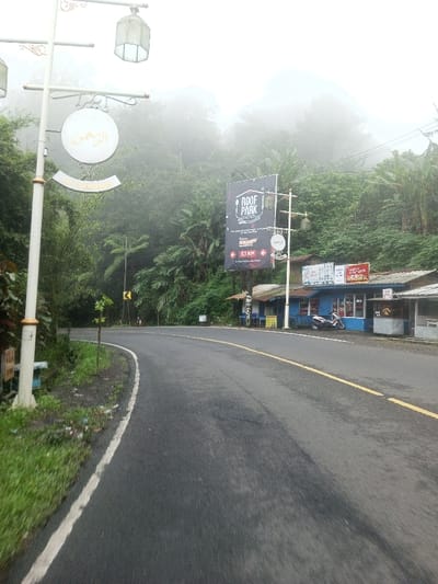 This image captures a curved, wet asphalt road in a misty, heavily vegetated mountainous area, with signage indicating a location near Pacet-Cangar, Indonesia, not Siruppiddy, Sri Lanka. The road features a solid white line along its left edge and a double yellow line towards the center. Dense green tropical foliage and trees line both sides, with fog obscuring the upper portions of the background vegetation.
In the left foreground, a white streetlamp pole with gold detailing supports two ornate lanterns and two white circular signs. The upper sign displays Arabic script, translating to "Ar-Rahman."
On the right side of the road, a large black vertical banner advertises "ROOF PARK KAFE & RESTO," specifying "One stop entertainment in Puncak Pacet Cangar" and indicating "5.1 KM" along "Jl. Raya Pacet Km. 5.1 Cangar." Beneath this sign, a row of small commercial buildings includes a prominent blue structure with various smaller signs affixed to its facade. A white scooter is parked in front of the blue building. Adjacent stalls with rustic, exposed timber frames are also visible.
Further along the left side of the road, a yellow diamond-shaped sign with a black arrow indicates a rightward curve. No people or animals are present in the scene. The sky is overcast, contributing to the soft, diffused lighting.