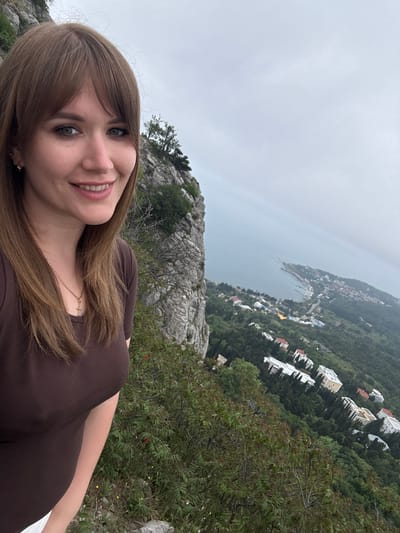 A woman stands on a rocky hillside overlooking the coastal town of Симеизский поселковый совет, Ukraine. The sea is visible in the background, along with buildings and greenery. The sky is overcast. The woman is wearing a brown top and a gold necklace. She has brown hair and is smiling.