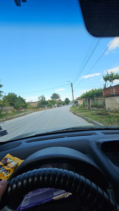 The image provides a driver's-eye view from inside a vehicle, looking out onto a narrow village road under a bright, clear blue sky with a few scattered clouds. The original image is rotated 90 degrees clockwise.
In the foreground, the dark grey dashboard and a black, ribbed steering wheel are visible on the right side of the frame. Tucked behind the steering wheel is a purple chocolate bar wrapper, partially revealing the words "Milk Chocolate" and a QR code. A yellow snack wrapper, possibly for "Lupu" branded cookies or biscuits, lies on the dashboard to the lower right. The upper left corner of the windshield shows a black, dotted frit band.
Outside the vehicle, a paved road stretches ahead, flanked by older, rustic houses on the left. These houses feature red tile roofs, worn stucco walls, and are often covered in green vines and foliage. A wooden utility pole with power lines and a street light stands prominently on the left side of the road. Abundant green trees and bushes line the street and surround the houses, suggesting a verdant, semi-rural setting consistent with Voiniagovo, Bulgaria.
The bright sunlight and clear sky indicate a sunny daytime, likely late morning or early afternoon. No people or animals are visible in the scene. The overall impression is one of a quiet, rural village road on a pleasant day.
