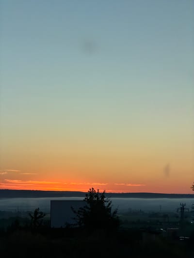 The image depicts a rural landscape at dawn or dusk, with a gradient of sky colors from pale blue at the top to vibrant orange and red near the horizon. A thick layer of fog or low-lying clouds obscures the ground level, creating a hazy effect over the distant landscape. Silhouetted against the colorful sky are rolling hills in the middle ground and a dark foreground with trees and what appears to be a large, blank screen, possibly part of an outdoor cinema or a billboard. Electrical poles and some structures are faintly visible through the fog in the distance. The scene suggests an outdoor setting, possibly on the outskirts of a town or village, given the faint signs of civilization. The overall atmosphere is tranquil and serene, bathed in the soft, diffused light of the setting or rising sun. The image does not contain any people or clear actions. Given the specific mention of Pașcani, Romania, this scene likely captures a view from a vantage point overlooking the surrounding countryside, with the fog being a common atmospheric phenomenon in that region during certain times of day and year.