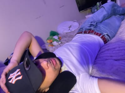 The image depicts an indoor scene in a room, purportedly located in Plaisir, France. A male individual is lying supine on a bed, angled diagonally from the lower-left to the upper-right of the frame. He is wearing a black New York Yankees baseball cap, a white short-sleeved t-shirt, light blue jeans, and a red studded belt. His left arm is bent with the hand near his head, which is tilted slightly to the right, revealing part of his face with eyes seemingly closed or averted, and lips slightly parted. The bed is covered with a light-colored, textured comforter, and a section of a purple blanket or pillow is visible on the right. In the background, on the bed, are several stuffed toys, including a white cat-shaped item and a green character resembling Yoshi. A flat-screen television is visible mounted on the wall in the upper-right background. The room is illuminated with a predominant purple-blue ambient light. The wall behind the subject's head is light-colored and shows minor dark marks. No other individuals are present, and no active interactions are depicted.