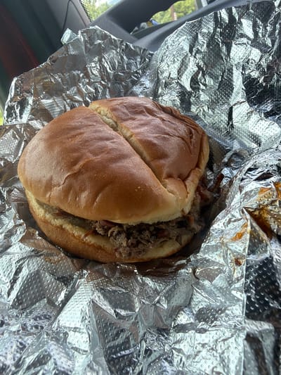 A close-up, overhead shot shows a pulled pork sandwich wrapped in crinkled aluminum foil. The sandwich is nestled within the foil, with the top bun slightly split. The pulled pork filling is visible between the bun halves, and some of the meat appears to have barbecue sauce on it. The foil has a textured, dimpled pattern. The background suggests the sandwich is inside a vehicle, with dark interior surfaces and a glimpse of green foliage and sky through a window. The image was taken in Chattanooga, United States of America.