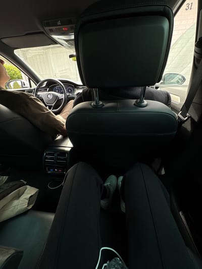 The image depicts the interior of a car, viewed from the back seat. The perspective is focused on the legs of two individuals in the rear, with one person's legs extended and another pair of legs partially visible. In the foreground, dark-colored trousers are prominent. White sneakers are worn by the individual whose legs are more visible. To the left, a beige garment is placed on the seat.
The front of the car is visible, including the dashboard, steering wheel with a Volkswagen logo, and part of the driver's arm. The driver is wearing a long-sleeved shirt in a muted green color. The car's interior features black leather seats. Above the dashboard, a control panel with buttons, including a red SOS button, is present.
The car's windows offer a view of an outdoor environment. The exterior appears to be a street scene with buildings and trees. One window shows a brick building with the number "31" visible. The scene suggests a bright day. The location is identified as Pori, Finland.