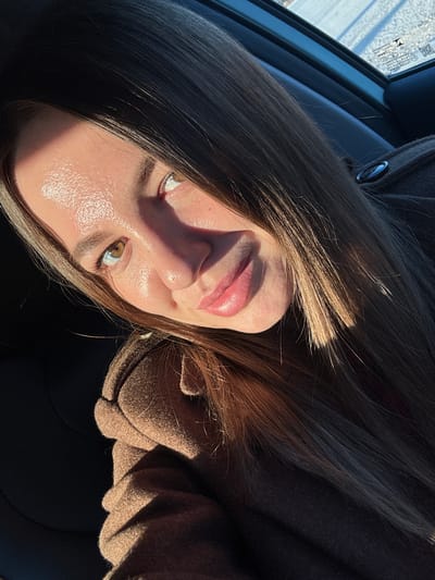 A selfie photograph shows a woman with light brown hair and fair skin, illuminated by sunlight. Her face is partially in shadow. She has hazel eyes and her lips appear to have a slight gloss. She is wearing a brown coat or jacket. The scene is inside a vehicle, as evidenced by the car window and interior elements visible in the background. The location is unknown and cannot be determined as Likino-Dulyovo, Russia from this image.
