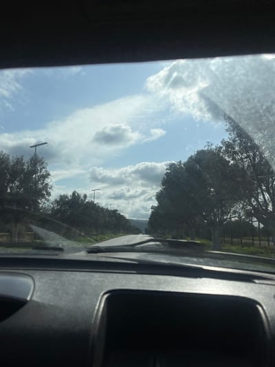 The image presents a view from inside a vehicle, looking forward through a windshield. The bottom quarter of the frame is occupied by a dark, textured dashboard. Two windshield wipers are visible at the base of the windshield. The windshield itself shows evidence of streaks and dust, with a significant glare from direct sunlight originating from the upper right quadrant.
A paved, two-lane road, appearing dry and without visible lane markings, extends into the distance. The road is bordered on both sides by dense rows of medium-sized trees, characterized by dark green foliage, consistent with evergreen or olive varieties. Modern, dark-colored, T-shaped streetlights are positioned at regular intervals along the road, receding into the background. A dark metal fence is partially visible behind the trees on the right side of the road.
In the background, distant hills or mountains are discernible beneath a sky featuring a mixture of bright blue patches and scattered white and grey cumulus clouds. The strong sunlight casts shadows and creates highlights on the clouds. The scene depicts a sunny day on a tree-lined thoroughfare within the city of Ain El Turk, Algeria. No individuals or other active vehicles are visible.