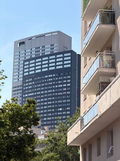 The image shows a street view in Courbevoie, France, on a clear day. In the foreground, a modern apartment building with beige walls and multiple balconies with glass railings is prominent. Some balconies have plants, furniture, and storage containers visible.

Behind the apartment building, a tall, dark gray skyscraper dominates the skyline. This building has a grid-like pattern of numerous windows and a lighter gray section at the top with a distinctive circular cutout feature. To the left of the dark skyscraper, another lighter gray building with a similar window pattern is visible.

Green trees with lush foliage are situated in the mid-ground and frame the view of the buildings. The sky is a bright, clear blue, indicating sunny weather. The scene captures an urban environment characterized by contemporary architecture.