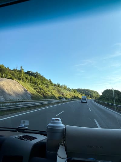 The image is taken from inside a vehicle, looking forward through the windshield at a highway. The sky is clear and blue. On the right side of the highway, there are trees and foliage covering rolling hills. On the left side, a steep, rocky embankment is visible, also covered with greenery. Several vehicles are on the highway, including a white van and a few cars. In the foreground, on the dashboard, a white insulated cup and a light-colored purse are visible. The image is consistent with a scene on a highway in a rural or semi-rural area, such as the outskirts of Malo Buchino, Bulgaria, during daylight hours.