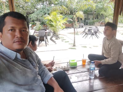 This image captures a relaxed outdoor scene in Keudealue Ie Puteh, Indonesia, likely at a cafe, restaurant, or resort. The perspective is a close-up selfie or a shot taken from very close to the foreground subject.
In the immediate foreground, on the left, a man with short dark hair and a mustache looks directly at the camera with a neutral expression. He is wearing a light gray patterned long-sleeved shirt and is seated on a wooden floor. Partially obscured to his right, another individual's hand is visible, holding a thin black object, possibly a pen or stylus. On the wooden surface in front of them are a white power strip with multiple sockets, a green tissue box, and a glass of iced tea with a straw. A plastic bottle with the word "AQUA" clearly visible on its label also sits on the floor.
To the right, a boy or young man sits cross-legged on the wooden floor, looking upwards and to his right with a contemplative or focused expression. He has dark, spiky hair and is wearing a light brown long-sleeved shirt and dark pants.
The background reveals a spacious open-air area under a partial thatched or wooden roof structure. Numerous outdoor dining tables and dark brown rattan-style chairs are arranged across a light-colored ground surface. The environment is lush with tropical foliage, including various trees, palm-like plants, and shrubs. Several wooden benches and distant buildings are also visible. The bright, even lighting suggests it is daytime, likely a sunny morning or afternoon. The overall atmosphere is one of leisure and tranquility.