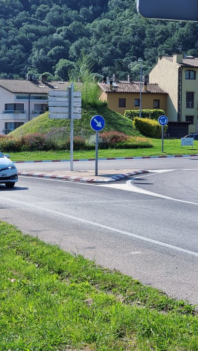 The image shows a street in Sant Joan les Fonts, Spain, with a roundabout ahead. A white car is visible on the left side of the image, partially obscuring a green lawn. The roundabout has a central island with a mound of grass and flowers, and a traffic sign indicating a mandatory turn to the left. Another sign indicates a roundabout. The buildings in the background are typical of Spanish architecture, with terracotta roofs and light-colored facades. The sky is blue and the sun is shining, indicating a clear day. The scene suggests a quiet, residential area with good road infrastructure.