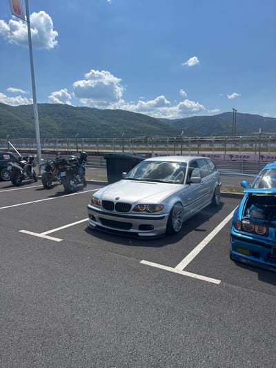 The image depicts a parking lot at a race track, likely in or near Dospey, Bulgaria, given the context of the race track. The scene is set under a bright, sunny sky with scattered white clouds. In the foreground, asphalt is visible with white parking lines. A silver BMW station wagon is parked in a marked spot, facing towards the right of the frame. It features custom headlights with colored halos. To the right of the silver BMW, a blue car with its hood open is partially visible.

In the background, several motorcycles are parked in a row. Behind the vehicles, there is a fence with various sponsor banners, including "DIANA". Beyond the fence, rolling green hills and mountains form the backdrop. A tall, silver pole with a flag is also visible on the left side of the image. The overall environment suggests a car or motorcycle event at a race track.