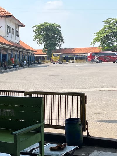 A bus station in Jati Wetan, Indonesia, is captured on a sunny day. The scene is dominated by a paved area with interlocking stones. To the left, a white building with orange-tiled roofs lines the station, featuring a row of arches and windows. In front of this building, several motorcycles are parked, and a few people are visible, some sitting and others standing near a red trash bin.

Further into the station, a bright yellow bus is parked on the left, and a red and white bus, marked with the number "2.0" and the logo "S", is parked on the right. The red bus has "PHANTIKA" written on its side. Signs are visible above the area where the buses are parked, with some text obscured but "AREA SHANT" discernable. Lush green trees are scattered throughout the background, suggesting a somewhat tropical environment.

In the foreground, a green wooden bench is partially visible on the left, with "JAYA" stenciled on its back. To the right of the bench, a dark blue barrel stands next to a dark metal fence with vertical bars. On a surface in front of the bench, a brown object, possibly a piece of fabric or a small toy, and a white object with some writing are present. The overall impression is one of a functional, open-air transportation hub under a clear blue sky.