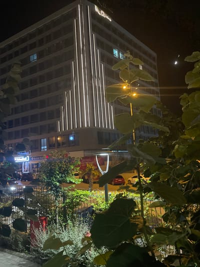 The image depicts a nighttime scene in the city of Yunus Emre, Turkey. A modern, multi-story building with vertical white lights illuminating its facade dominates the background. The building has numerous windows, some of which are illuminated from within. A sign with a logo and text, possibly a business name, is visible at the top of the building.
In the foreground, lush green foliage partially obscures the view of the building and the street below. Several large leaves are prominent in the lower portion of the image. Beyond the foliage, a fence separates the foreground from a sidewalk and street. Parked cars, including a red and a white vehicle, are visible on the street.
Additional elements include streetlights casting a yellow glow and two crescent moons visible in the dark night sky. Signs for businesses, including "Teknoloci" and "Kazan Köfte," are discernible on the lower levels of the building. The overall impression is a well-lit urban environment at night, with a blend of modern architecture and natural elements. No people are directly visible in the scene.