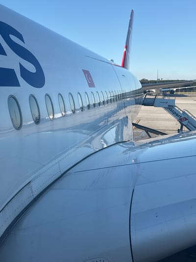 A wide-body Turkish Airlines aircraft is parked on an asphalt tarmac under a clear blue sky at Guarulhos, Brazil. The aircraft's white fuselage features a partial blue "S" graphic and a small rectangular Turkish flag emblem. A series of oval passenger windows extends along the fuselage, reflecting the sky. The aircraft's red vertical stabilizer is partially visible in the background. The top surface of the aircraft's right wing, characterized by metallic paneling and rivet lines, is prominent in the foreground. A white mobile passenger stairs unit, branded "Swissport" with the designation "SWB 0253," is positioned adjacent to the aircraft's fuselage. In the distance, the tail and wing of another large white aircraft are partially visible, with a line of green trees on the horizon. No individuals are depicted.