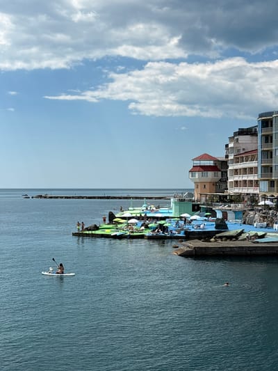 In Kiparisnoe, Ukraine, a picturesque coastal scene unfolds under a partly cloudy sky. The azure sea dominates the foreground, its surface rippling gently. A lone figure on a paddleboard, accompanied by a dog, glides across the water. In the mid-ground, a vibrant waterfront recreation area beckons with colorful inflatable structures, lounge chairs, and umbrellas. People are seen relaxing and enjoying the sun. The background is defined by a series of buildings lining the coast, their architecture reflecting a mix of styles. Further out, a long pier extends into the sea, with a few distant figures visible. The overall atmosphere is one of leisure and seaside tranquility.