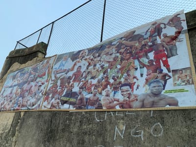 The image shows a concrete wall covered in posters, located at a site in Sokoto, Nigeria with coordinates of 13.049 latitude and 5.238 longitude. The posters, which appear to be advertisements, feature a collage of images of men, some of whom are shirtless. The men are posing in various ways, possibly in boxing stances. The posters are partially obscured by a chain-link fence. There is a sign reading "Sokoto" on the lower part of one of the posters. On the wall below the posters, the word "NGO" is written in white chalk.