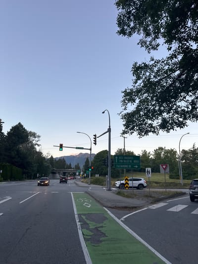 The image shows a street scene in North Vancouver, Canada, likely near an intersection. A road with multiple lanes is visible, with a dedicated green bike lane on the right side. Traffic lights are present, with a green light illuminated for one direction and a red light for another. Several cars are on the road, some in motion and others stopped.
Overhead signs indicate directions to "WEST VANCOUVER", "LYNN VALLEY RD. NORTH", and "2nd NARROWS BR.". Pedestrian crossing signs and a yield sign are also visible. Trees line the sides of the road, and mountains are visible in the background, suggesting a scenic location. The sky is clear with a pale blue hue, indicating it is either early morning or late evening. The overall impression is of a typical urban intersection in a city with natural surroundings.