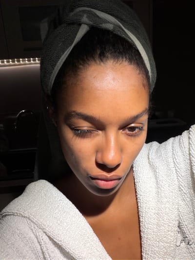 A close-up, indoor portrait shows a young woman of color with dark hair wrapped in a green towel, wearing a white terrycloth robe. She is winking with her right eye closed and her left eye open, looking slightly down. Natural light illuminates her face from the right, creating strong shadows on the left side. The background is dark and out of focus, but suggests a domestic setting with kitchen elements like a faucet and under-cabinet lighting visible on the left. The scene appears to be in Champs-sur-Marne, France.