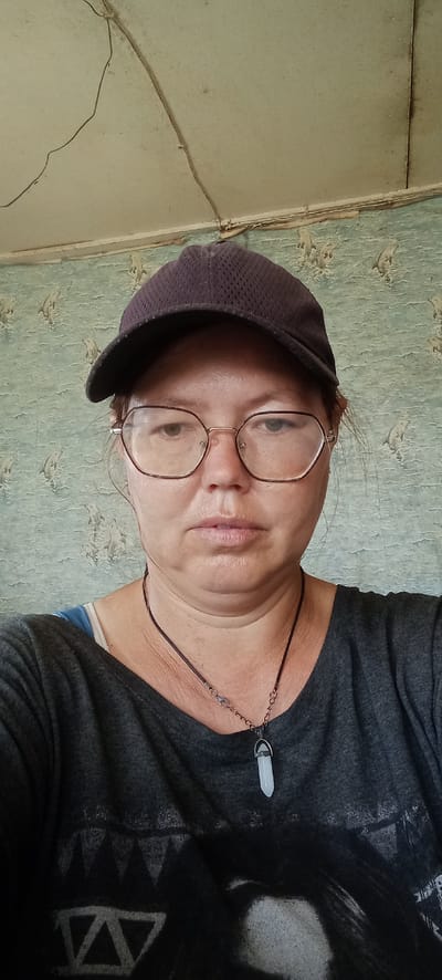 A woman wearing a dark baseball cap and eyeglasses is looking at the camera. She is wearing a dark grey t-shirt and a necklace with a light-colored pendant. The background consists of a wall with light blue wallpaper featuring a repeating pattern of what appear to be leaping dolphins, and a discolored ceiling with visible cracks and wires. The overall impression is of an indoor setting, possibly a residential interior. The location in Январцев ауылдық округі, Kazakhstan is not discernible from the image content.