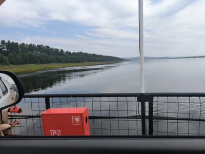 The image shows a view from a ferry crossing the Angara River in Boguchany, Russia. In the foreground, a red fire extinguisher box is visible on the deck of the ferry, secured by a black metal fence. The box has some Cyrillic lettering and a symbol on it. To the left, a portion of a white vehicle, likely a car, is reflected in a side mirror. The river stretches out to the horizon, with a lush green forest lining the opposite bank. The sky is partly cloudy, with a serene atmosphere. The ferry appears to be moving, as indicated by the gentle ripples on the water.