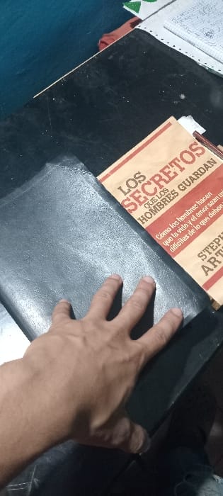The image captures a close-up, slightly overhead view of a person's right hand resting on a dark surface, possibly a desk or table. The hand, with its palm facing down and fingers slightly spread, is positioned over a rectangular, dark gray or black object that appears to be a book or a flat folder, possibly made of a slightly textured material like leather or a similar synthetic.

To the upper left of the hand, partially visible, is a brown-covered book or publication, tilted diagonally. The cover displays text in Spanish. Prominently visible are the words "LOS SECRETOS QUE LOS HOMBRES GUARDAN" (THE SECRETS THAT MEN KEEP) in large, bold letters. Below this, smaller text reads: "Cómo los hombres hacen que la vida y el amor sean dificiles de lo que deben" (How men make life and love more difficult than they should be). Further down, the partial words "STEP" and "AR" are visible, likely part of an author's name or a series title. The word "RDAN" is also visible at an angle, part of the larger "GUARDAN" text.

The primary surface supporting these items is dark, possibly black, and shows some light specks or dust. On the far left, a white piece of paper with some faint markings or drawings is partially visible, along with a hint of green and a reddish-brown object underneath it. A dark blue or turquoise surface is visible on the bottom left edge of the frame. The right side of the image is dominated by shadows and a dark, indistinct object. The lighting appears somewhat muted, suggesting an indoor setting with possibly ambient or artificial light.