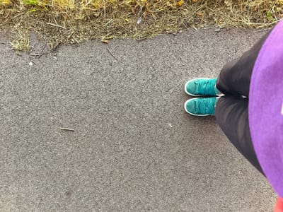 The image is an overhead shot of a person's legs and feet standing on a paved surface. The person is wearing teal colored sneakers and dark pants. A purple shirt is visible on the right side of the frame. The paved surface appears to be asphalt and borders a strip of dry, yellowish grass. A small twig lies on the asphalt in the lower left quadrant of the image. The lighting suggests it is daytime. This scene is located in Wervershoof, Netherlands.