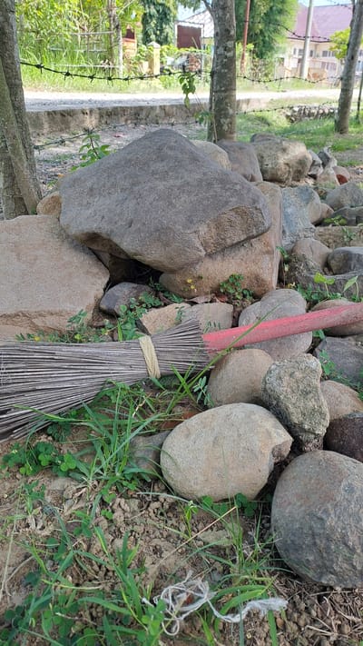 The image depicts a pile of large, weathered stones situated in an outdoor environment. A broom, its bristles made of dry, thin stalks and bound with twine, lies horizontally across some of the rocks, with a faded red handle extending to the right. Green grass and small, clover-like plants grow among the stones and on the ground. In the background, a barbed wire fence runs horizontally across the scene. Beyond the fence, a dirt path is visible, leading to a building with a white and pink roof and what appears to be a courtyard with some greenery and trees. The overall scene suggests an outdoor, possibly rural or semi-rural, area, with elements of both nature and human-made structures. No people are present in the image. The location is identified as Payalipah, Indonesia.