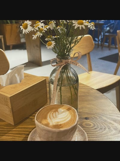The image depicts a close-up of a table setting, likely in a cafe or restaurant. A wooden tissue box with a white tissue peeking out sits on the left. To its right, a glass vase filled with water contains a bouquet of small white daisies. A light-colored ribbon is tied around the neck of the vase. In the foreground, a cup of latte with latte art is placed on a saucer. The table surface is made of weathered, light-colored wood. In the background, out-of-focus chairs and furniture suggest an indoor setting. The scene does not provide any direct information about the city of Saint-Août, France, nor does it contain any people or actions.